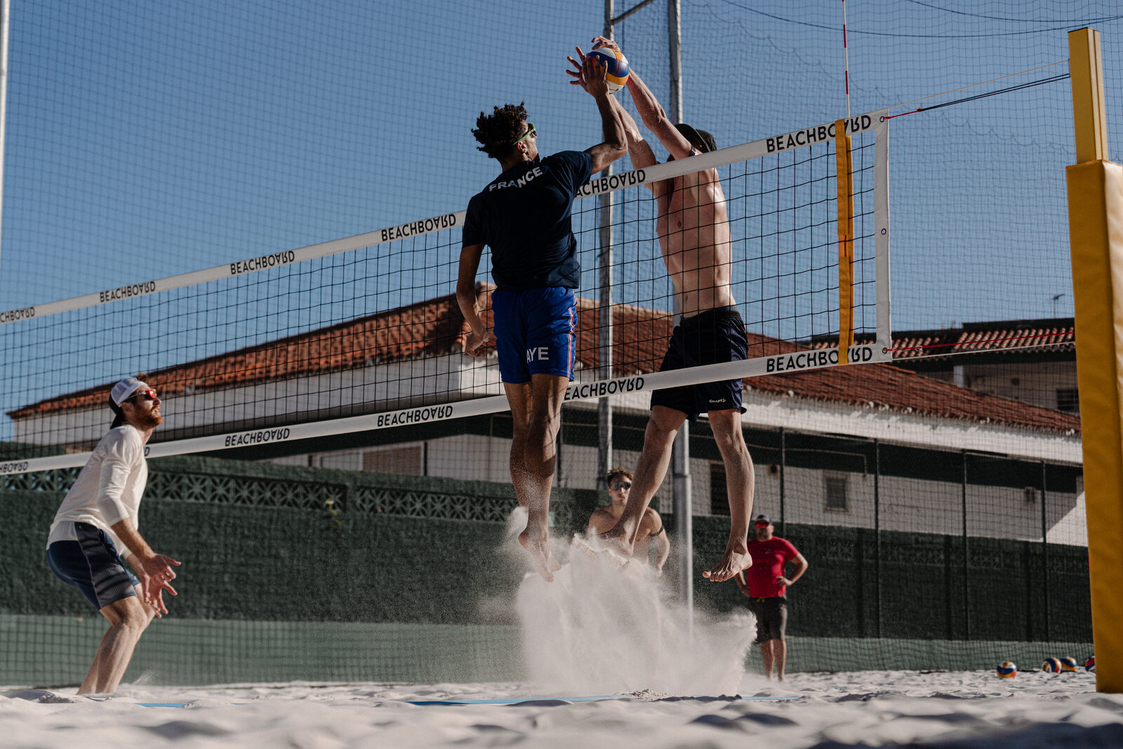 Two players block together at the net, sand kicking up on a Hacienda court